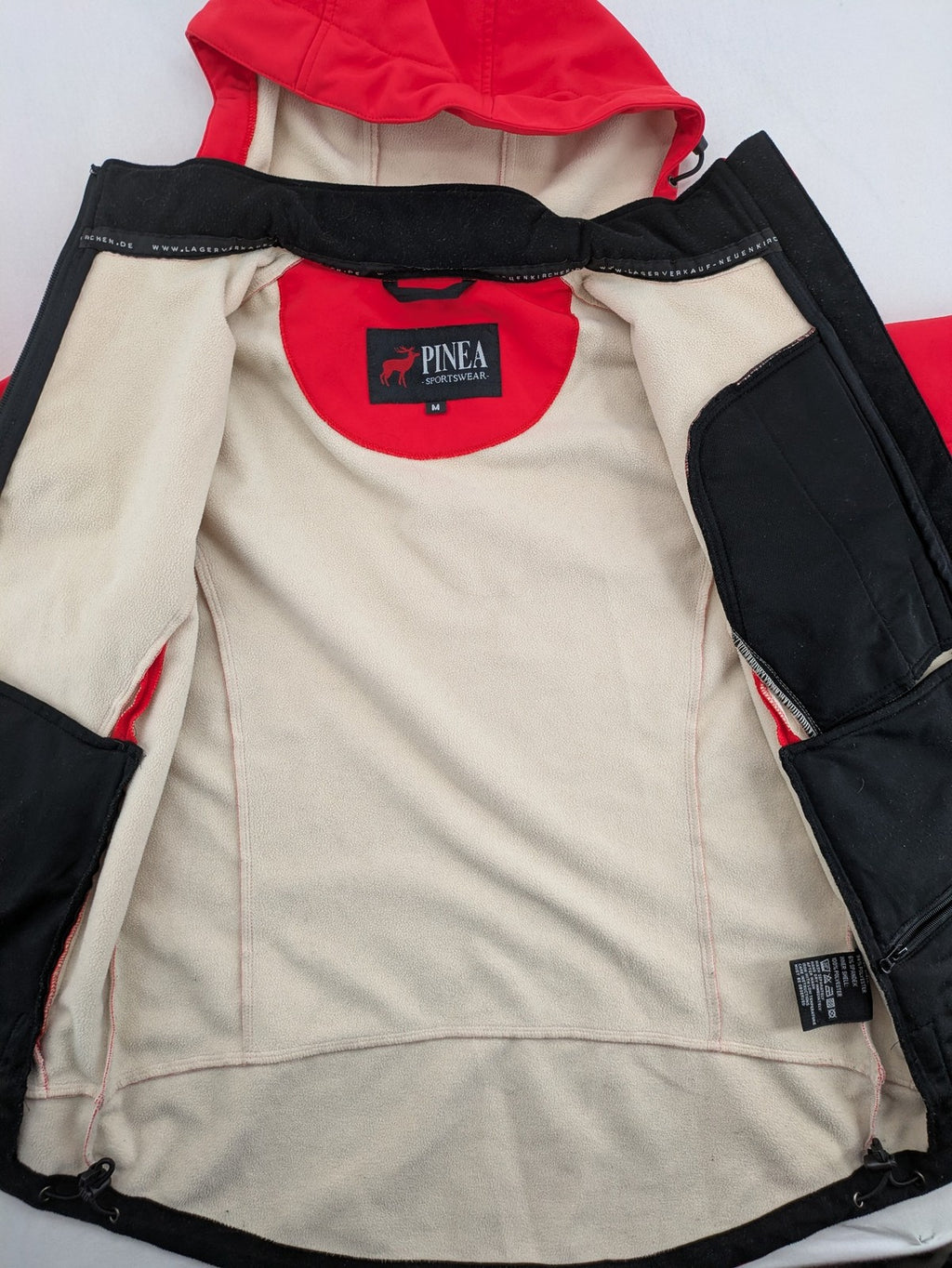 Pinea red softshell jacket with adjustable hood and fleece lining, multiple zippered pockets.