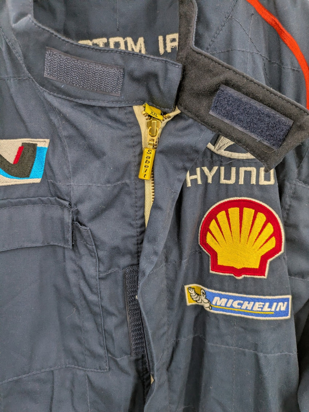 Hyundai Motorsport Sabelt racing suit navy blue with embroidered Shell and Michelin logos.