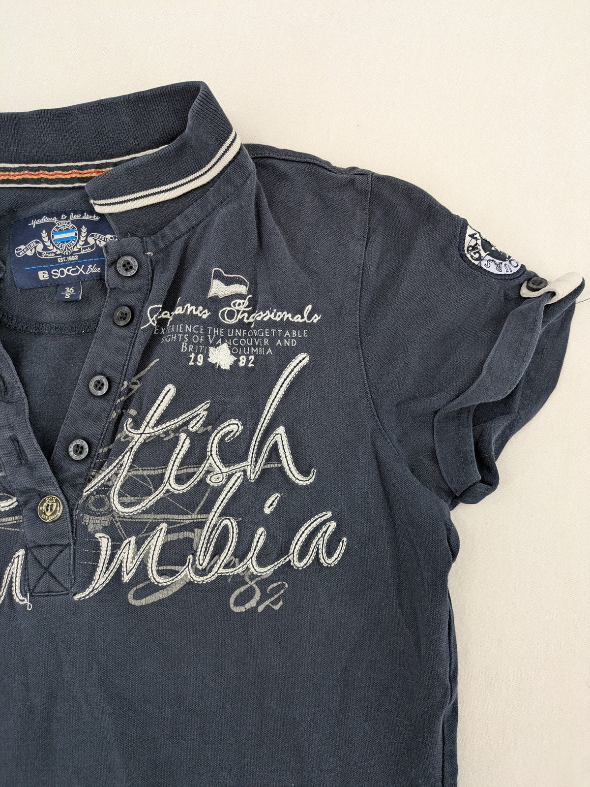 Soccx blue polo shirt with British Columbia graphic, embroidered patches, and striped collar.