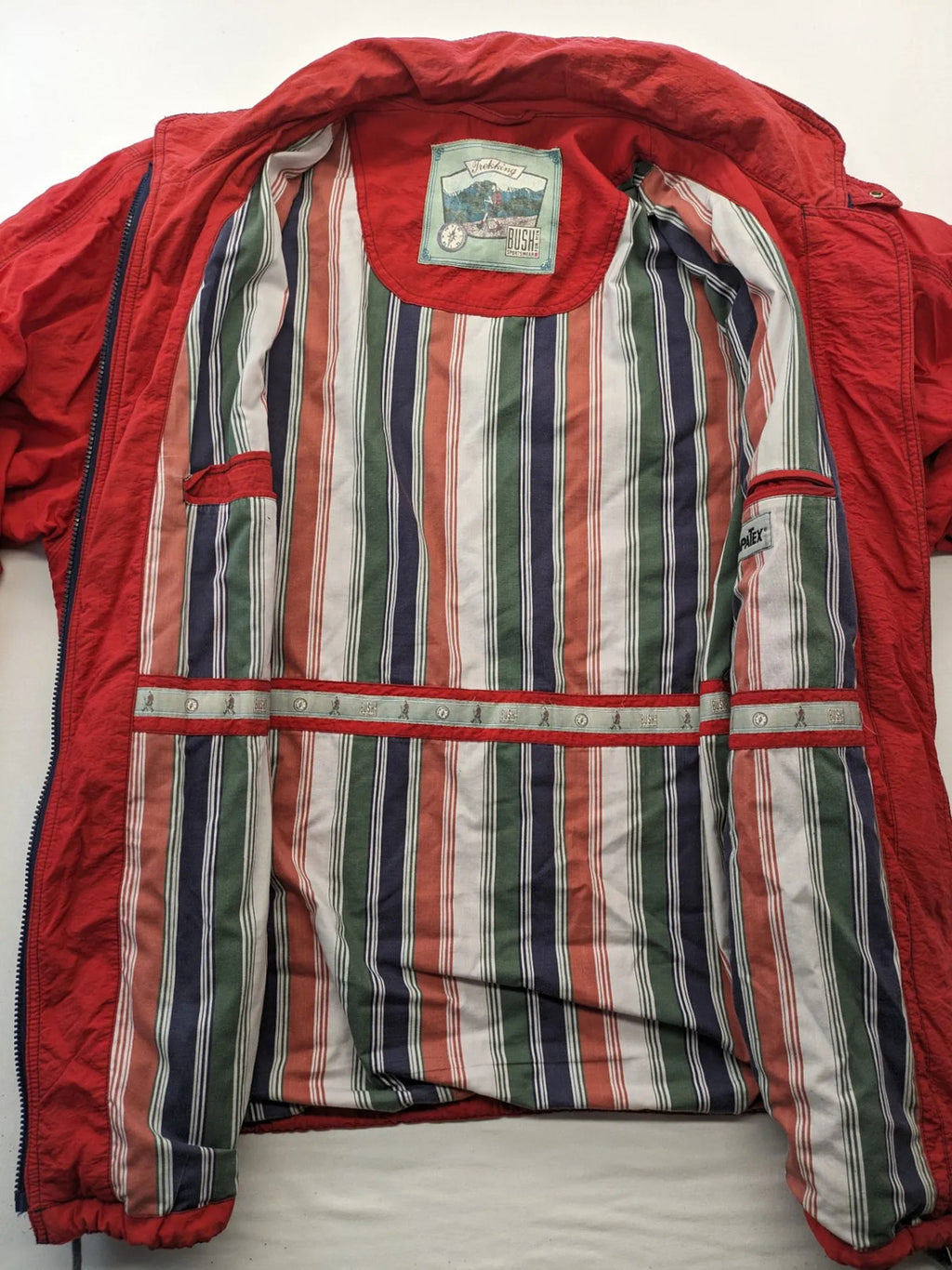 Bush Sportswear red trekking jacket with front zipper, multiple pockets, and striped lining.