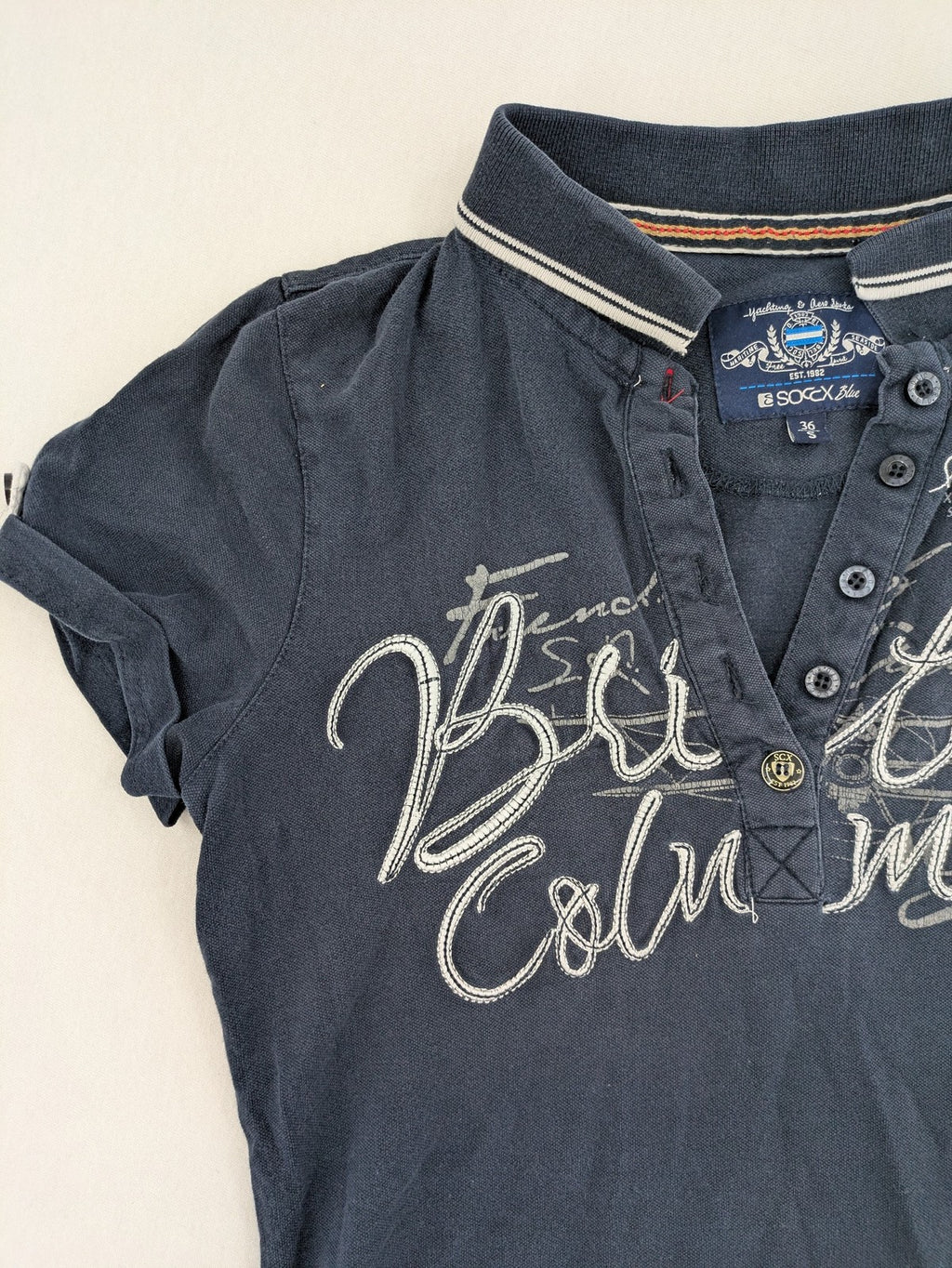Soccx blue polo shirt with British Columbia graphic, embroidered patches, and striped collar.