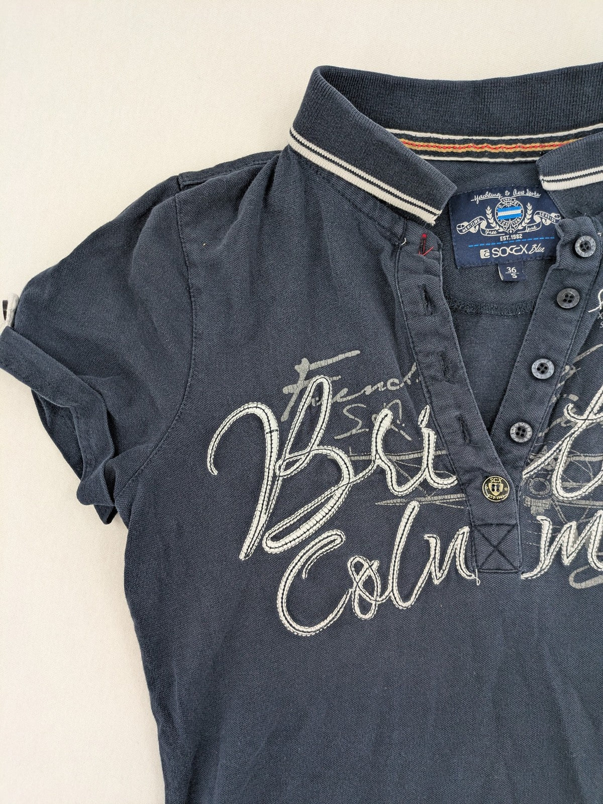 Soccx blue polo shirt with British Columbia graphic, embroidered patches, and striped collar.